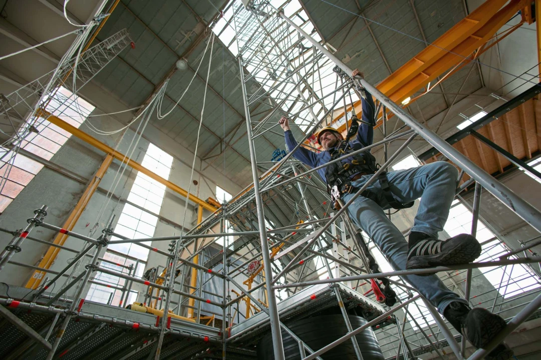 man die klimt op ladder in loods met helm op en harnas aan