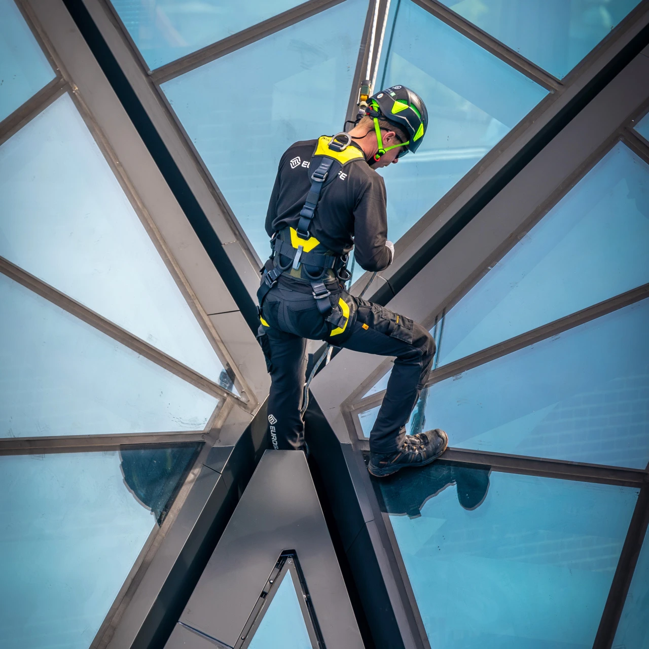 man in harnas met helm op aan touw vast staande op glazen dak
