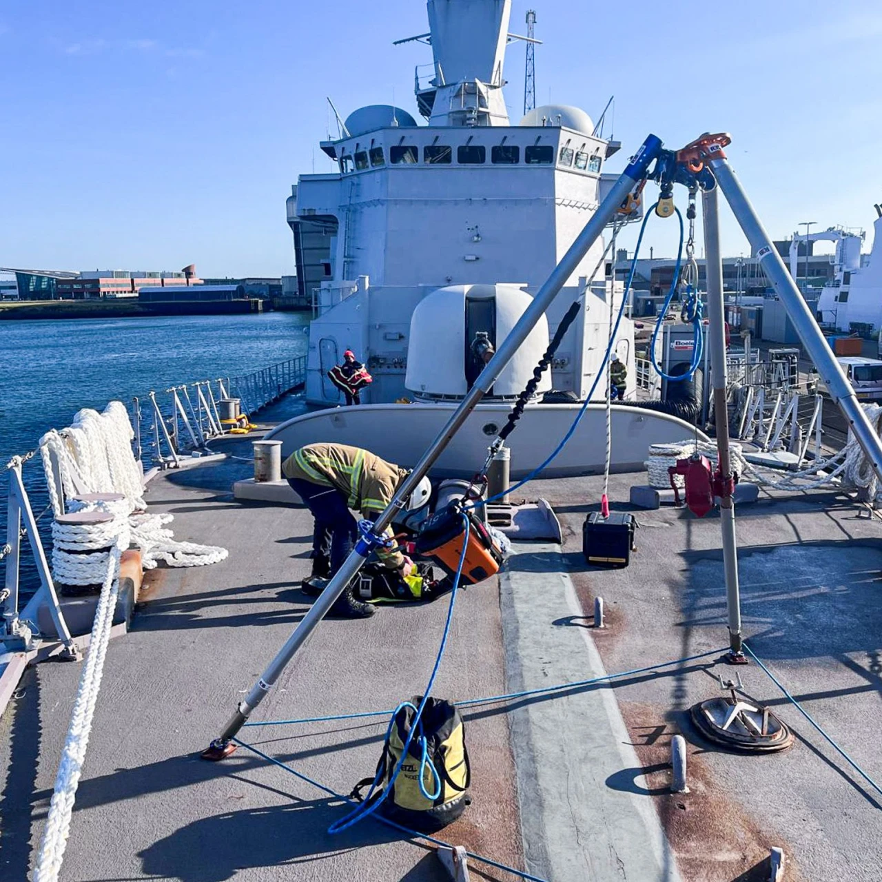 een groot schip waarbij een brandweerman naast een driepoot staat met touwen en een touwbrommer.