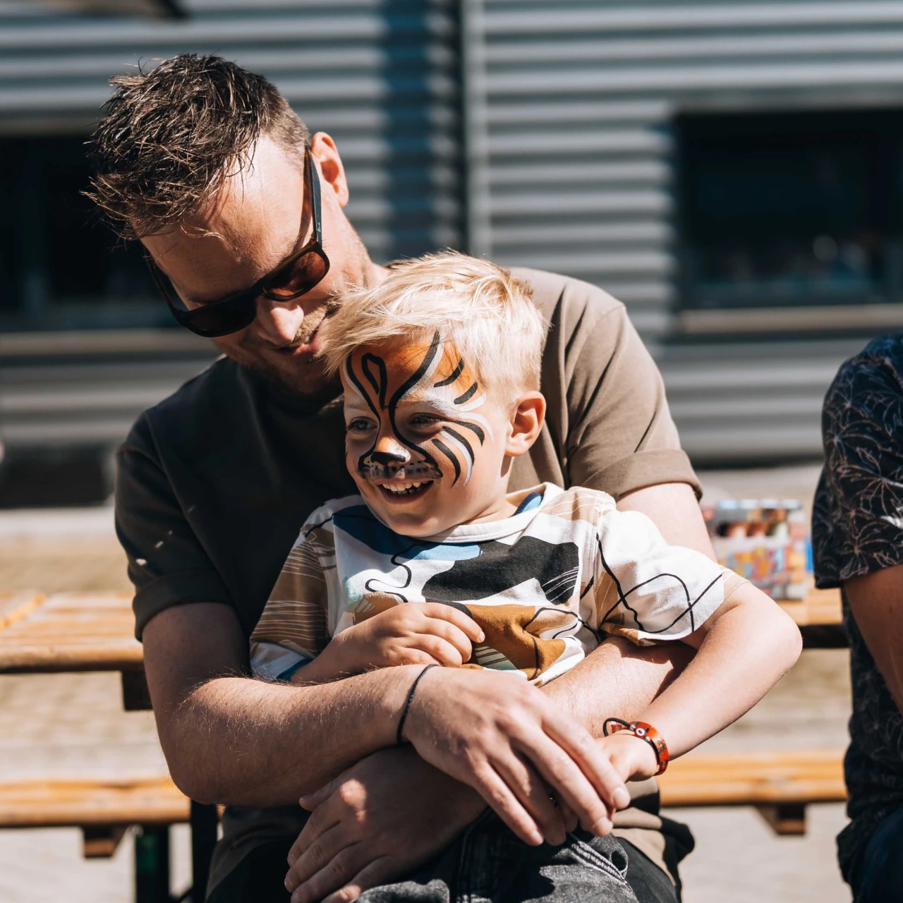 een man die een zonnebril draagt met een kind op schoot die schmink op zijn gezicht heeft van een leeuw