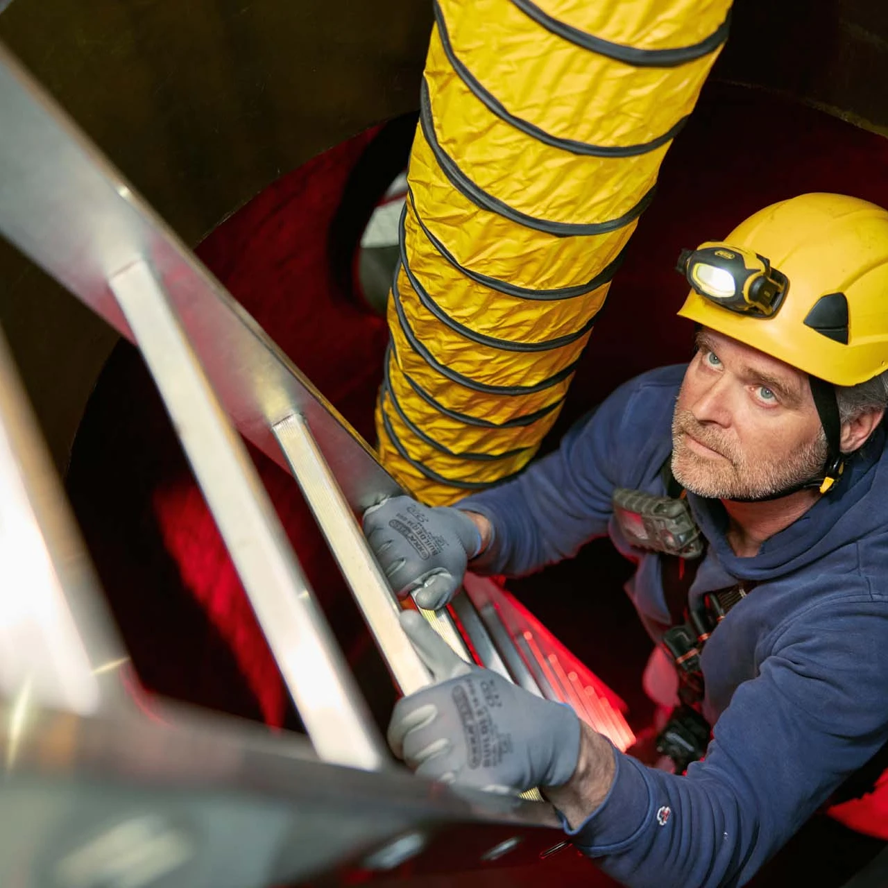 man op ladder in besloten ruimte met helm en lamp op hoofd