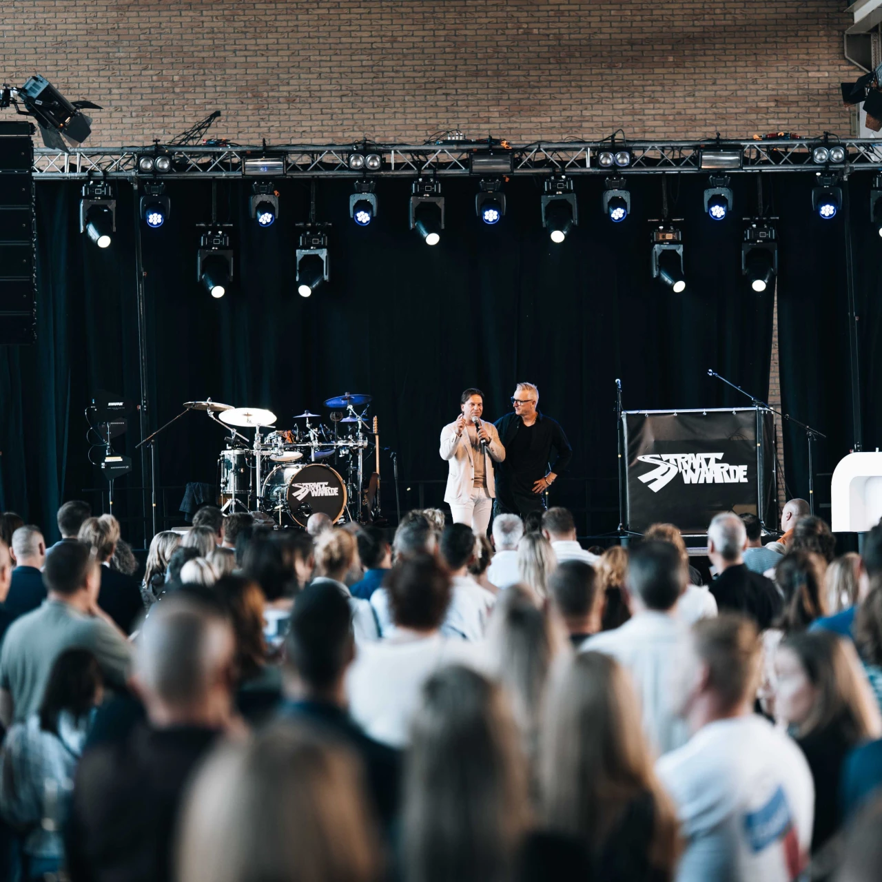 twee mannen die staan op een podium en een speech geven aan tientallen personen