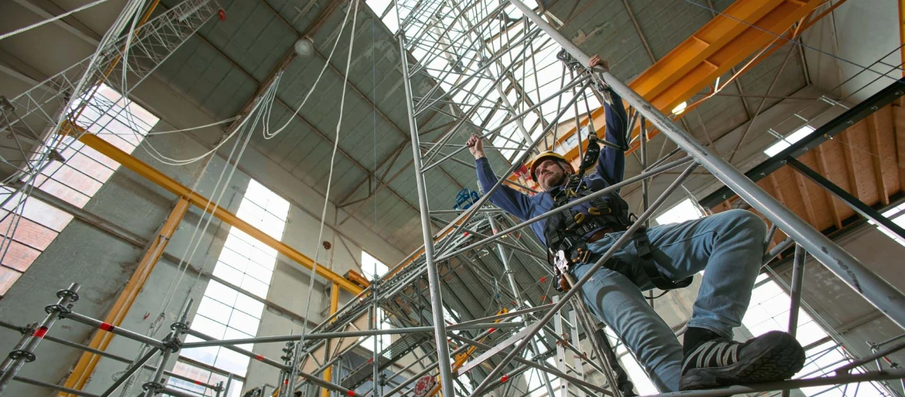 man die klimt op ladder in loods met helm op en harnas aan