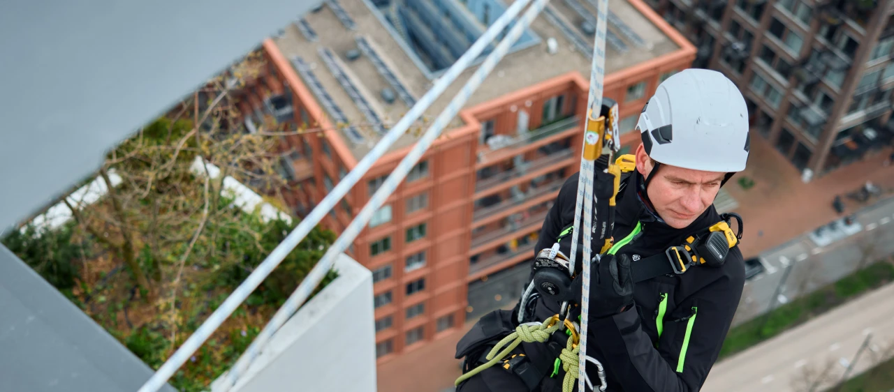 man hangend aan touw aan hoog gebouw in harnas en met helm op
