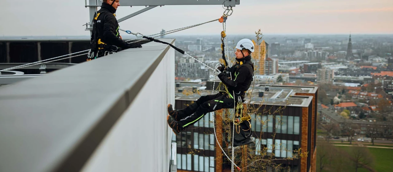 mannen op dak hangend aan touw in harnas met helm op