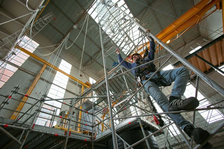 man die klimt op ladder in loods met helm op en harnas aan